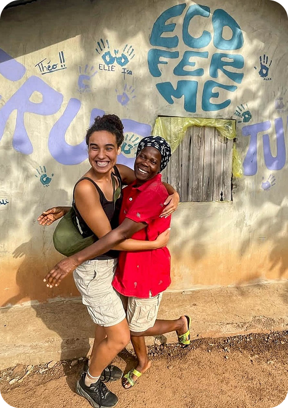Two women embracing in front of a wall with 'Eco Ferme' graffiti.
